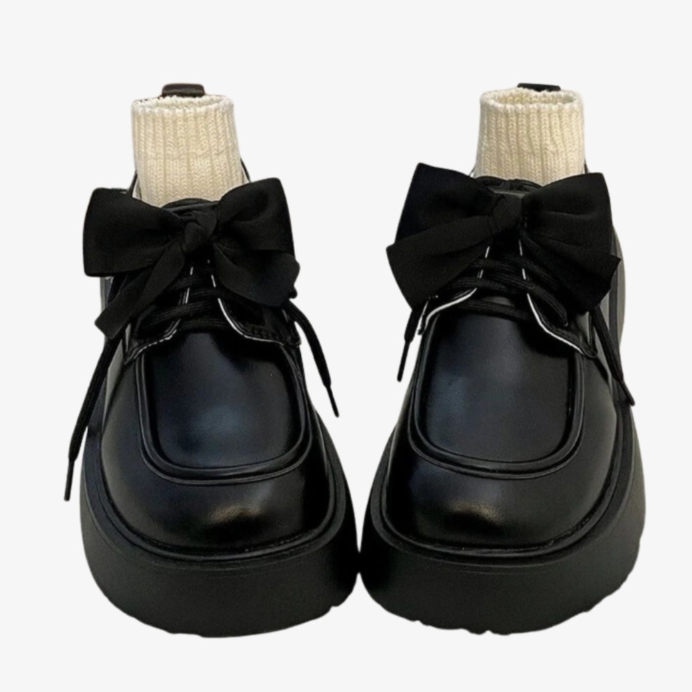 A pair of loafers in smooth black leather set on a white bed, a large bow decorates the top and little beige socks fill the shoe.
