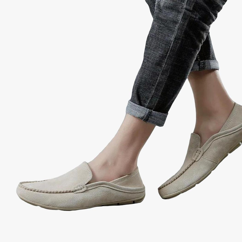 On a light gray background, the lower legs of a man in jeans and his beige genuine leather loafers