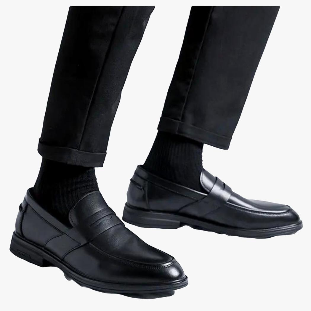 The lower legs of a man on a light background, wearing black pants and classic black faux leather loafers