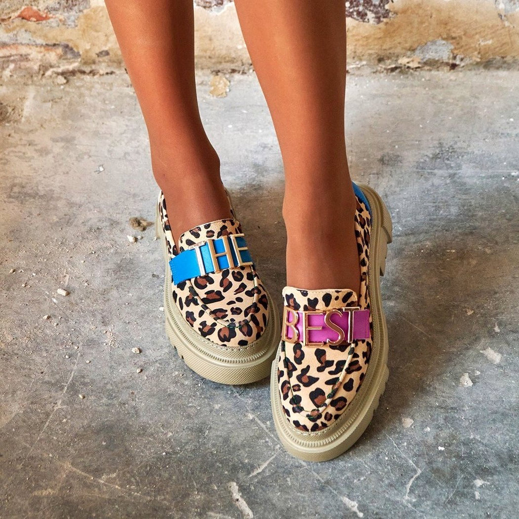 On an outdoor floor, a pair of loafers worn from the front in beige with a thick platform sole, leopard print with a blue strap over turquoise with gold lettering "THE" on one shoe and a pink strap with "BEST" on the other