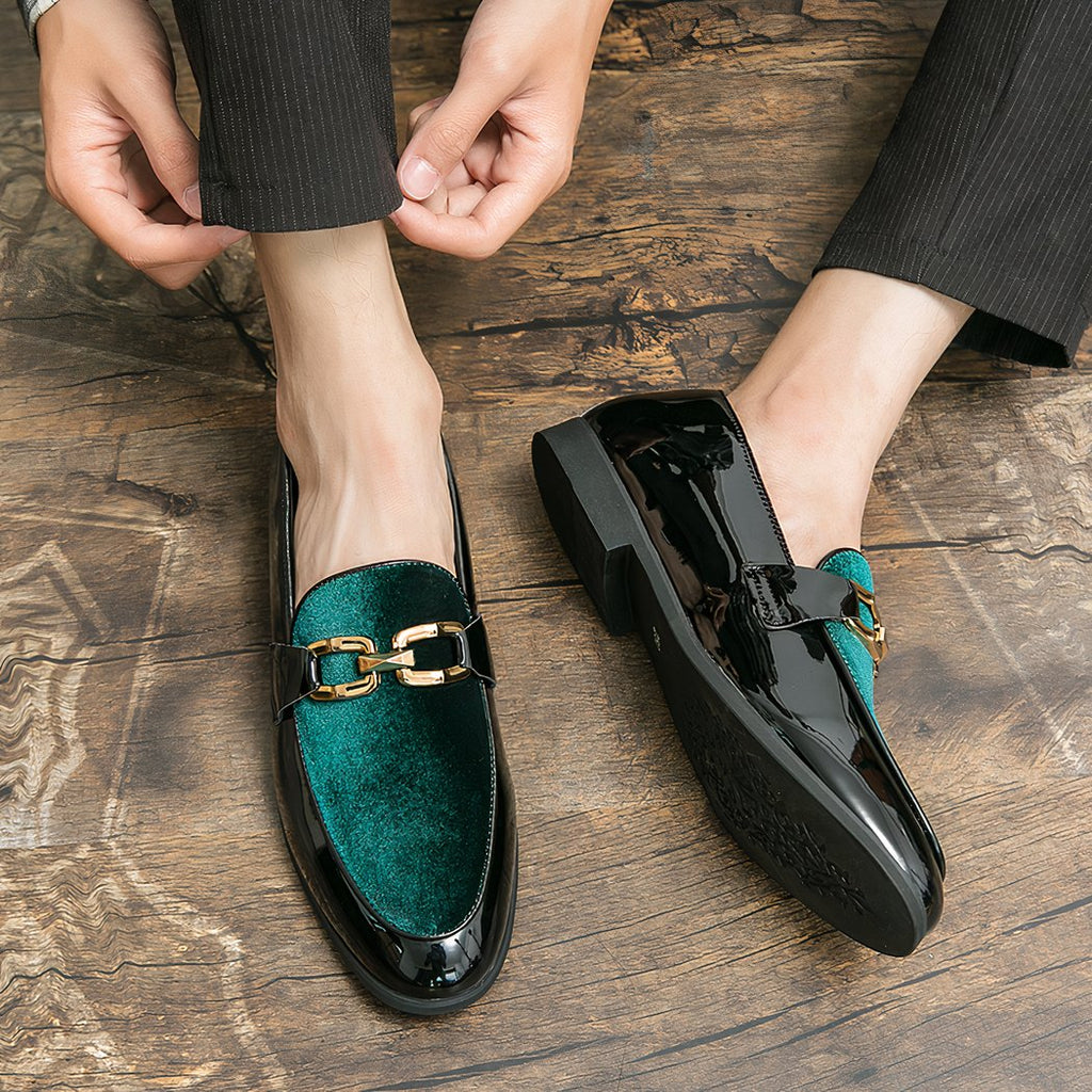 Pair of men's loafers in black patent on the turn and shiny green velvet with gold bit on the top. They are worn with black pants on a brown parquet floor