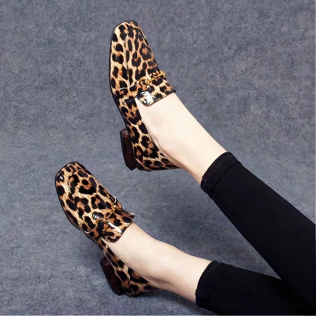 On a grey background, a pair of legs with black leggings and shiny loafers with leopard print square heels