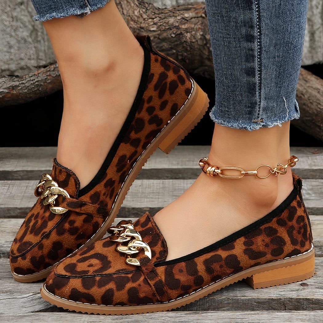 Worn with jeans and exposed ankles, we see in profile a pair of brown leopard print loafers with a small imitation wood heel and a gold chain decoration on top. There's a gold ankle chain on one foot.