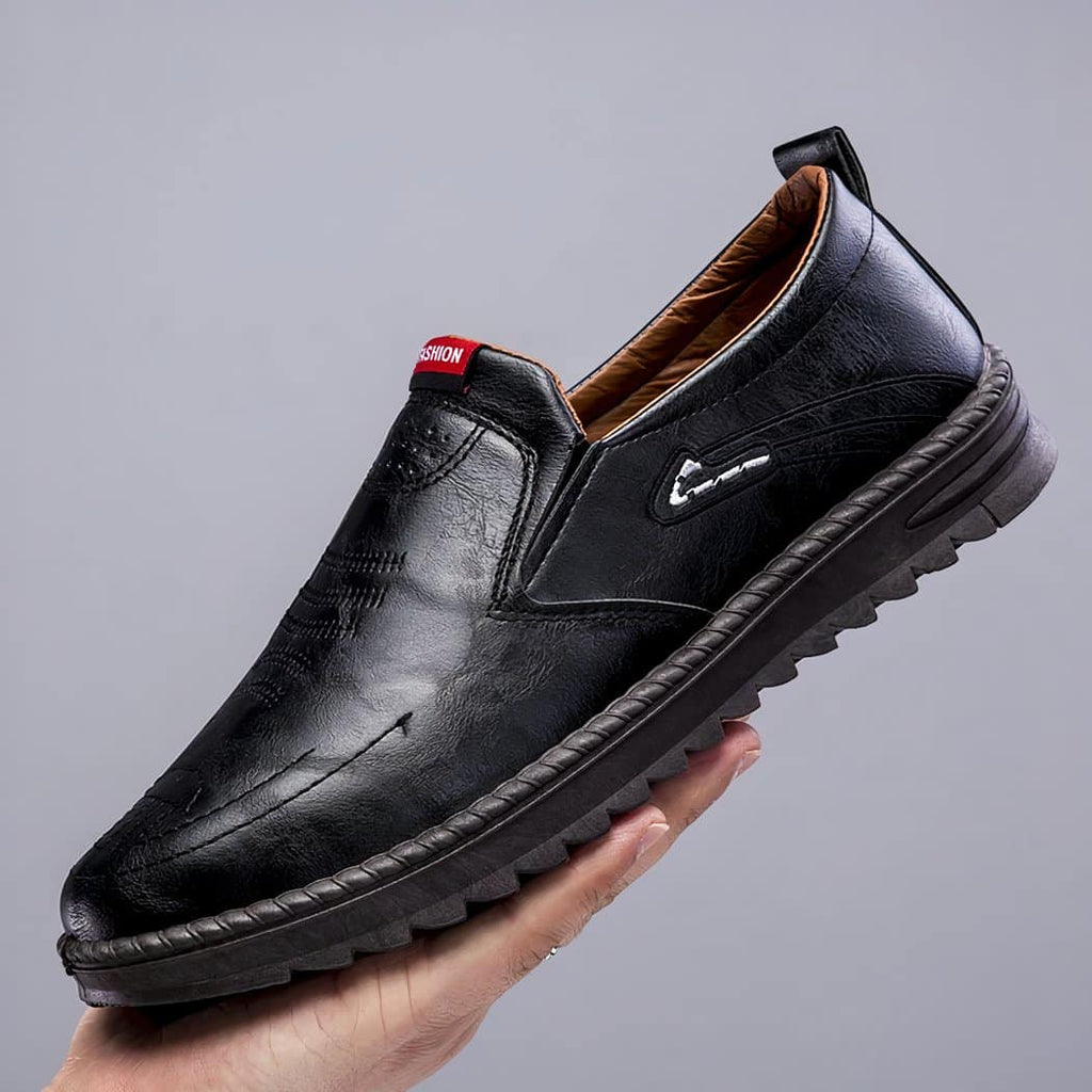 Black loafer with thick sole, held in one hand. Grey base.