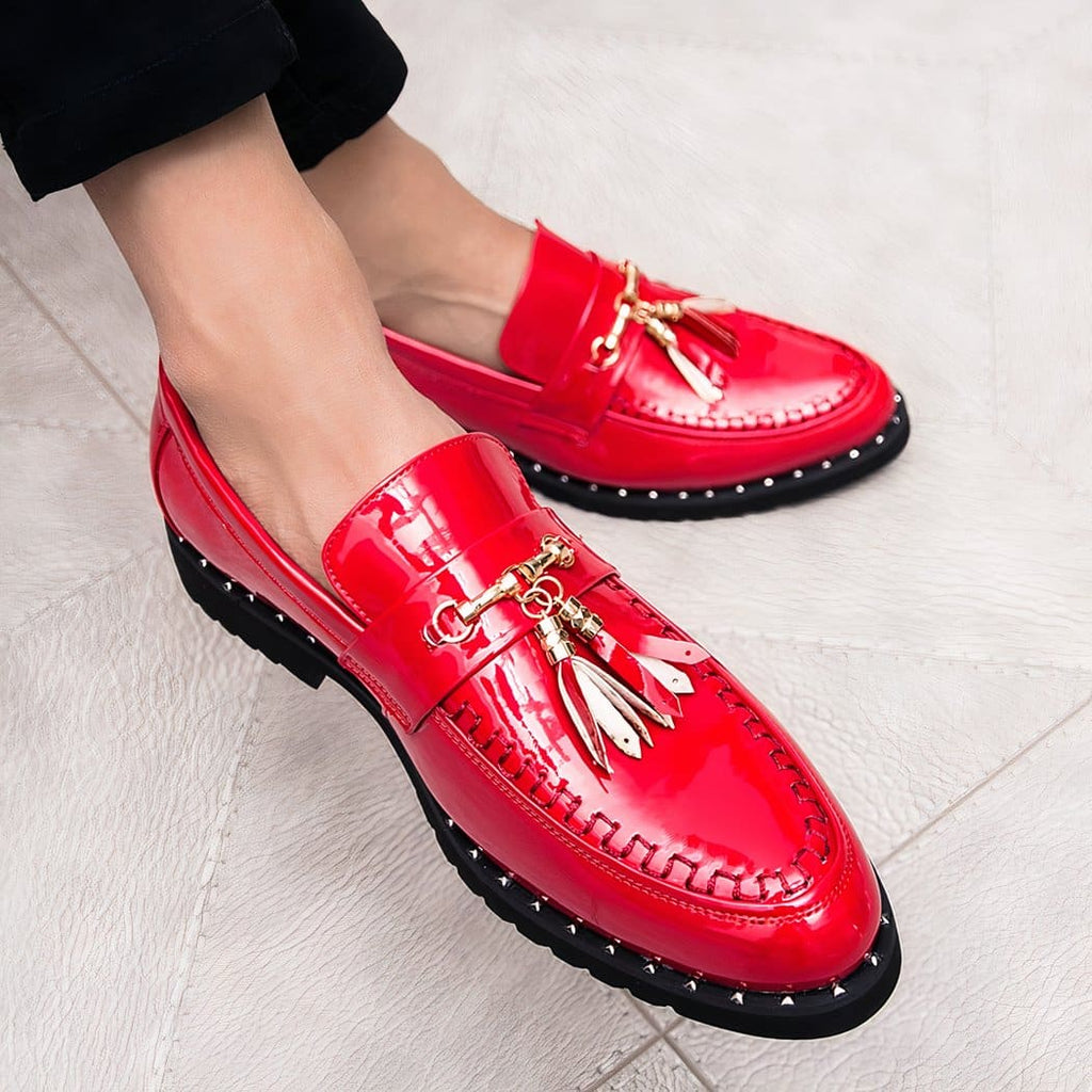 Bright red patent loafers with decorative tassels on the feet of a man sitting cross-legged in black suit pants.