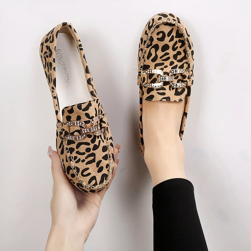 Pair of leopard-print loafers with stitching and silver rhinestone rings on top. One shoe in one foot and the other held in one hand.