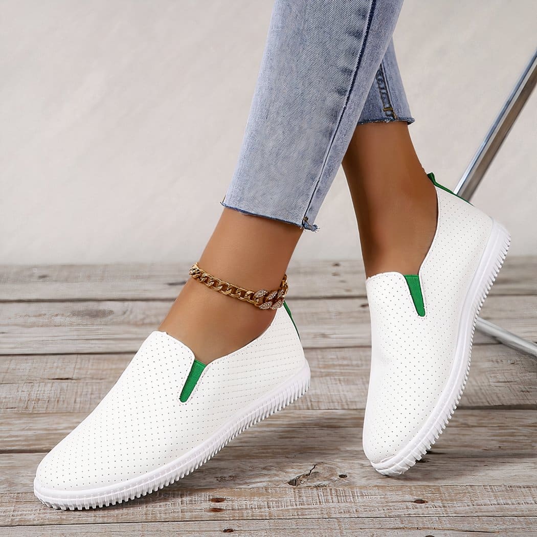 On a gray parquet floor, a pair of perforated white loafers with small green elastics on the sides. They are worn barefoot with light blue jeans and ankle chain.