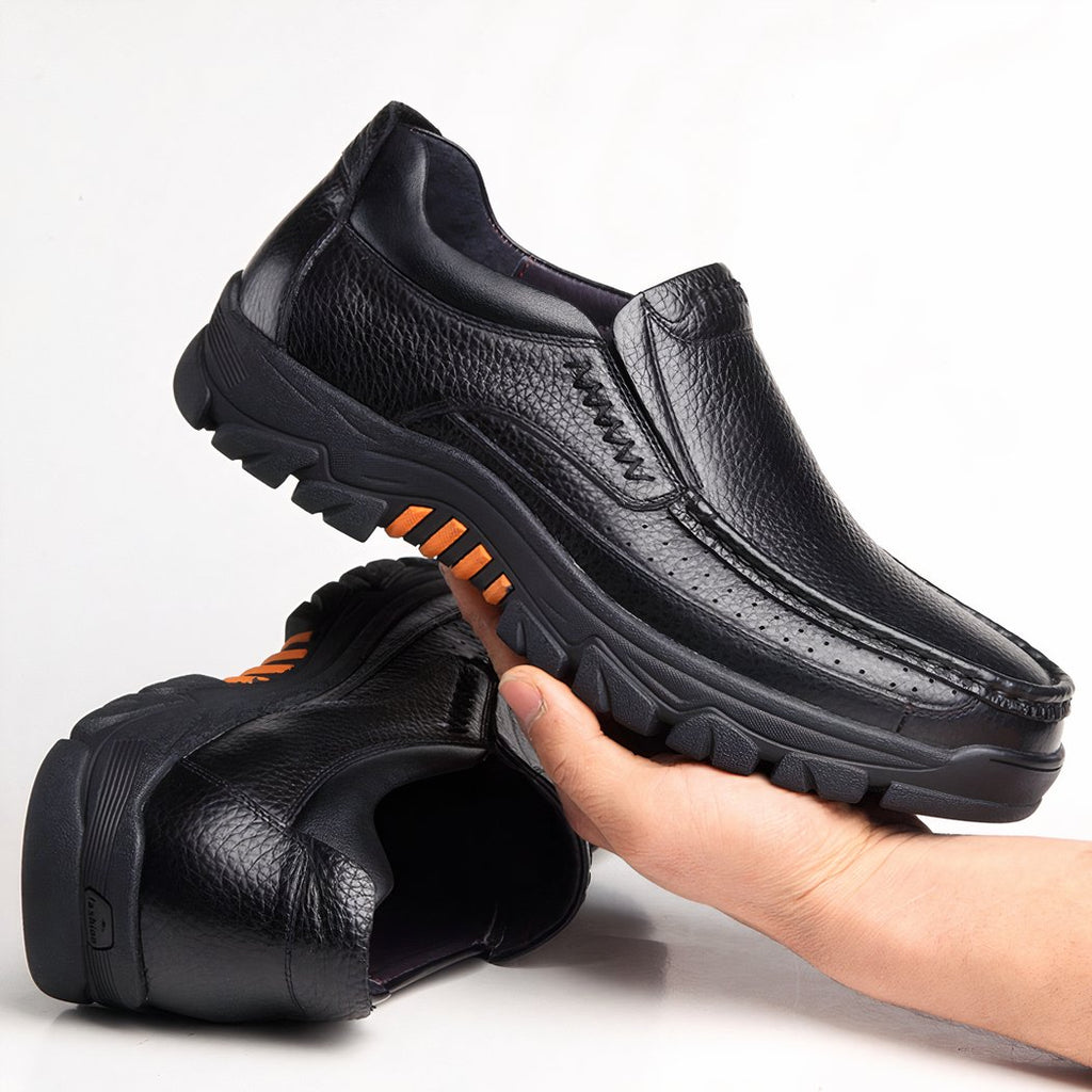 Photo of a pair of black leather orthopedic loafers with thick soles, one shoe lying on the floor, the other held in one hand.