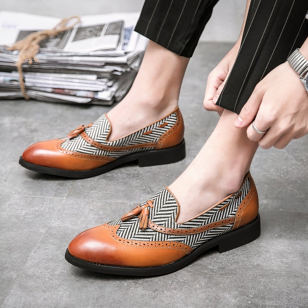 Two-material loafers with a white and black herringbone pattern and light brown leather and decorative tassels on the feet of a man wearing black suit pants.