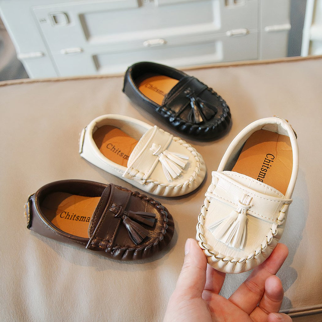 Several small tassel loafers for children on a table and one held in one hand.