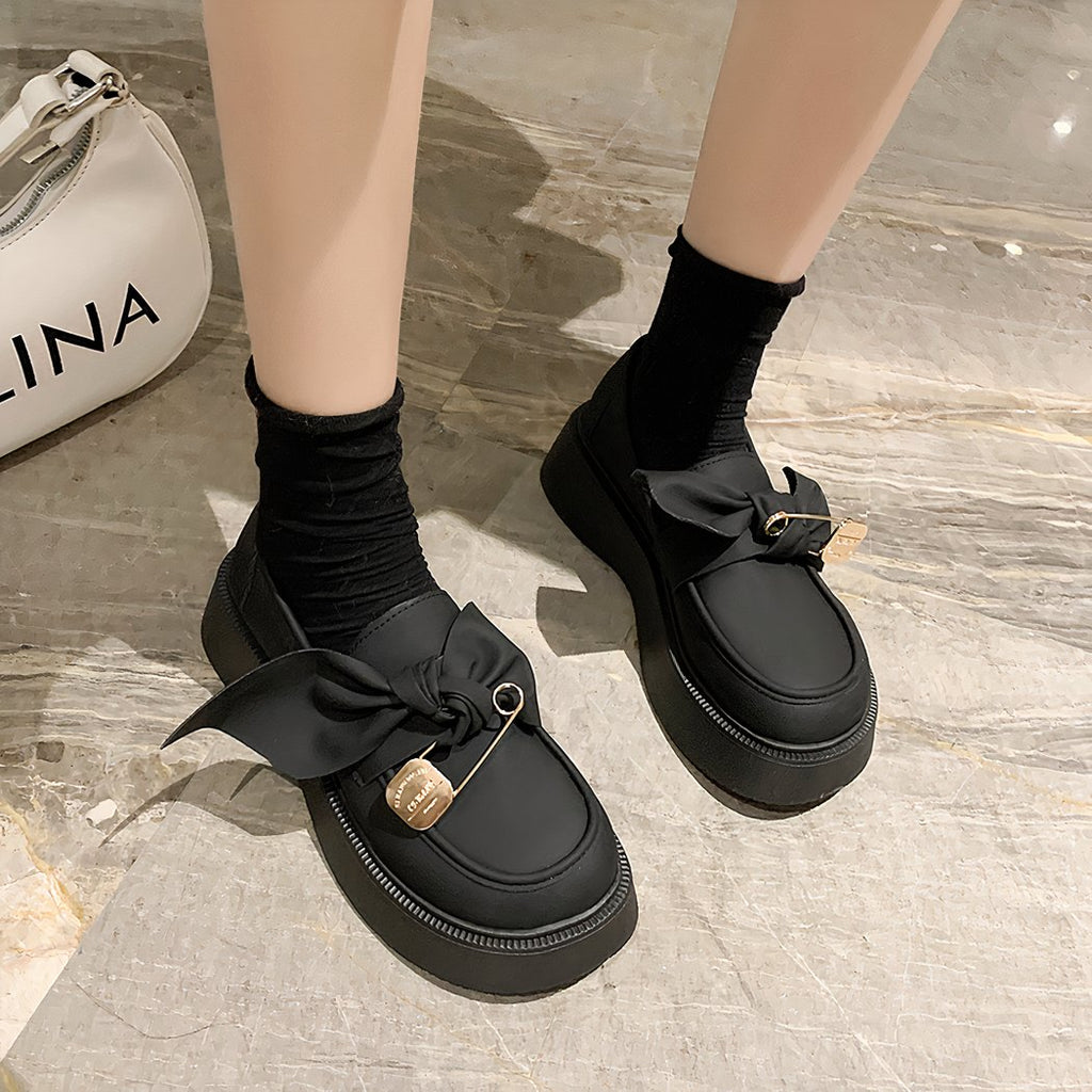 Round, smooth, thick-soled loafers with a big bow and a fake pin decorating the top of the shoe at the feet of a woman standing on a white marble floor and wearing little black socks.