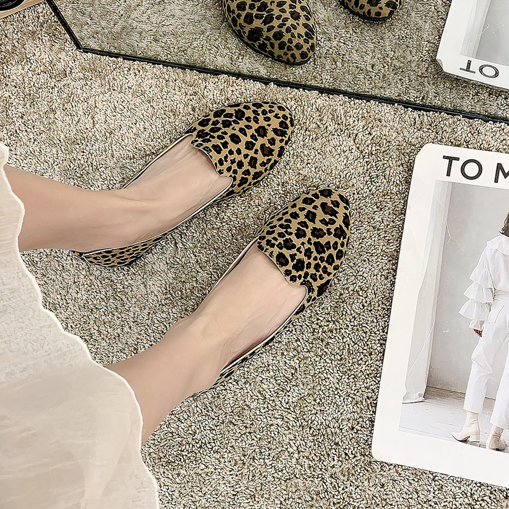 Pair of simple loafers with leopard print seen from above worn with a white skirt and bare feet. The floor is beige carpet and there's a white magazine next to it