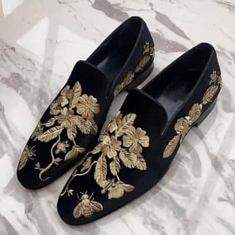 On a white marble surface, a pair of black velvet moccasins with gold floral embroidery
