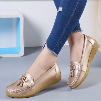Photo of a pair of gold moccasins worn by a woman with jeans, on a gray floor with a white wall and a white stool