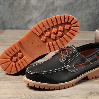 Photo of a pair of black boat moccasins with brown soles, on a parquet floor with a gray wall in the background