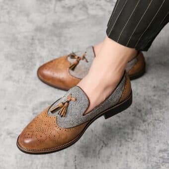 On gray ground, a pair of men's two-material dress loafers in marbled brown synthetic leather with stitching pattern and gray striped canvas with tassel on top.