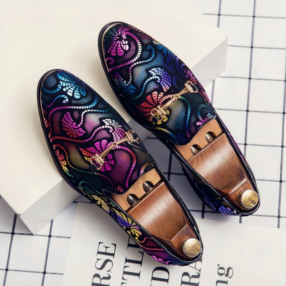 On a checkered surface with black and white lettering, a pair of evening moccasins with multicolored arabesques and silver bits. There are wooden shoe trees with a gold button inside the shoes.