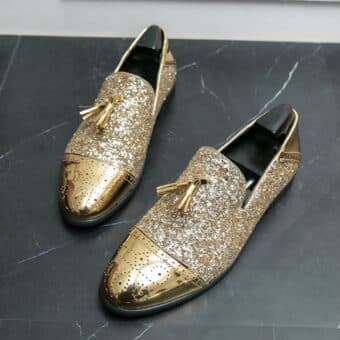 On a gray marbled floor, a pair of varnished and glittered gold moccasins with tassels and black shoe trees inside