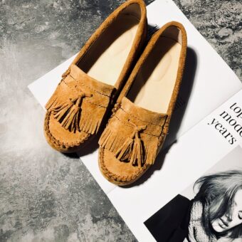 Indian-style camel suede loafers with fringe and tassels, set on a magazine on a waxed concrete floor.