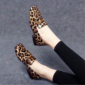 On a grey background, a pair of legs with black leggings and shiny moccasins with leopard print square heels