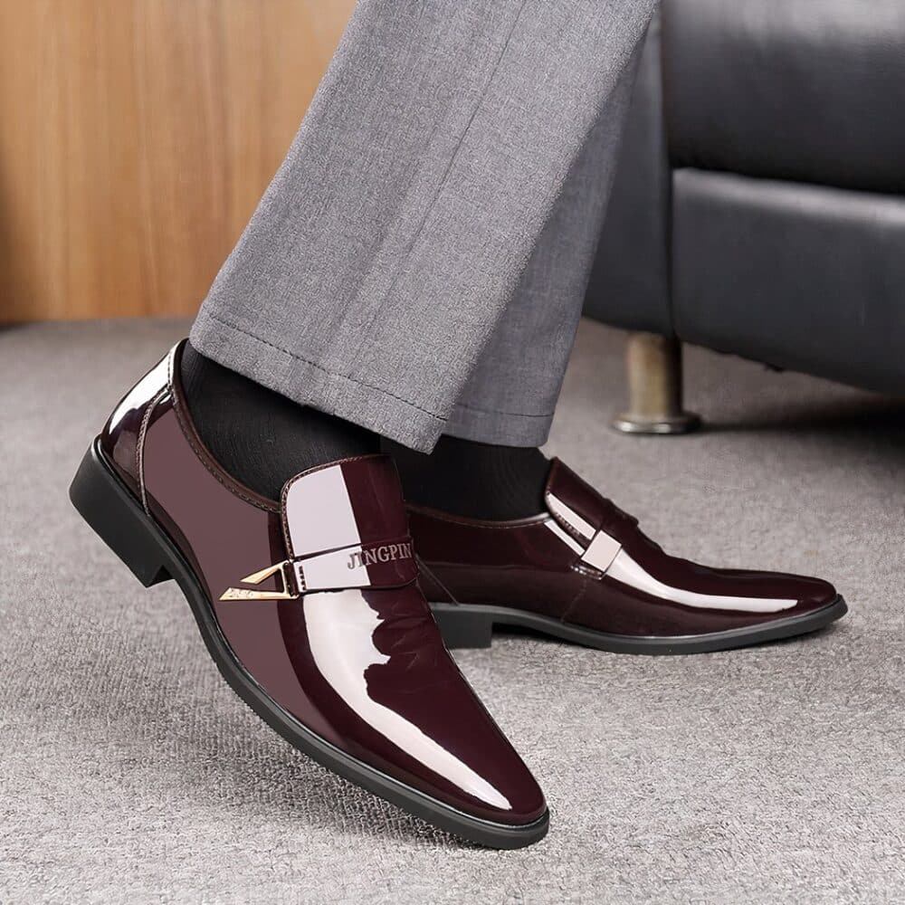 Feet in brown patent leather loafers and grey pants on a grey carpet