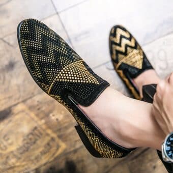 Worn barefoot, a pair of moccasins in black with gold rhinestone geometric embroidery. One foot rests on a brown parquet floor.