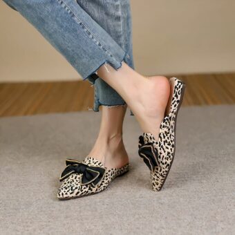 On a gray carpeted floor, a pair of pointed mule loafers with a black bow on top, a leopard print adorned with gold rivets. The pair is worn with short, ripped blue jeans, the ankles bare.