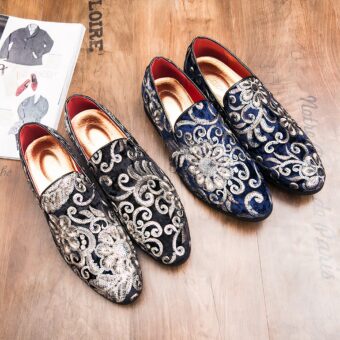 Two pairs of black and blue moccasins in shiny velvet with floral embroidery and sequins and a gold insole. They are set on a brown wooden surface next to a menswear magazine