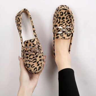 Pair of leopard-print moccasins with stitching and silver rhinestone rings on top. One shoe in one foot and the other held in one hand.