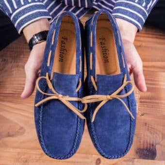 A pair of blue moccasins held in the hands of a person wearing a watch and a blue and white striped shirt