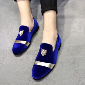 Pair of shiny blue velvet loafers with gold strap on front and embossed tiger head patch on top. Worn with black pants on a gray floor
