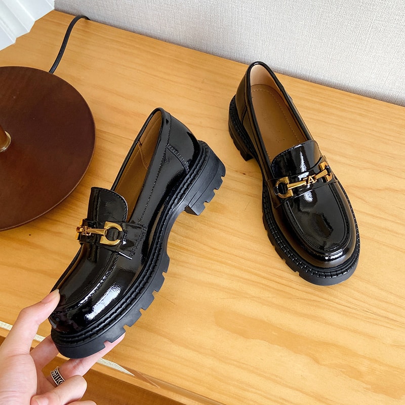 Photo of a pair of black patent leather moccasins held by someone, resting on a wooden cabinet next to a lamp