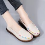 Photo of a pair of beige moccasins worn by a woman in black pants, against a gray background