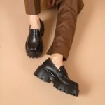 Photo of a pair of black moccasins worn by a woman with brown pants on a pink background