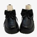 A pair of moccasins in smooth black leather set on a white bed, a large bow decorates the top and little beige socks fill the shoe.