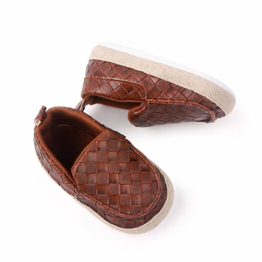 On a white background, a pair of brown square-toe moccasins with braided leather straps on the top and beige rope around the shoe