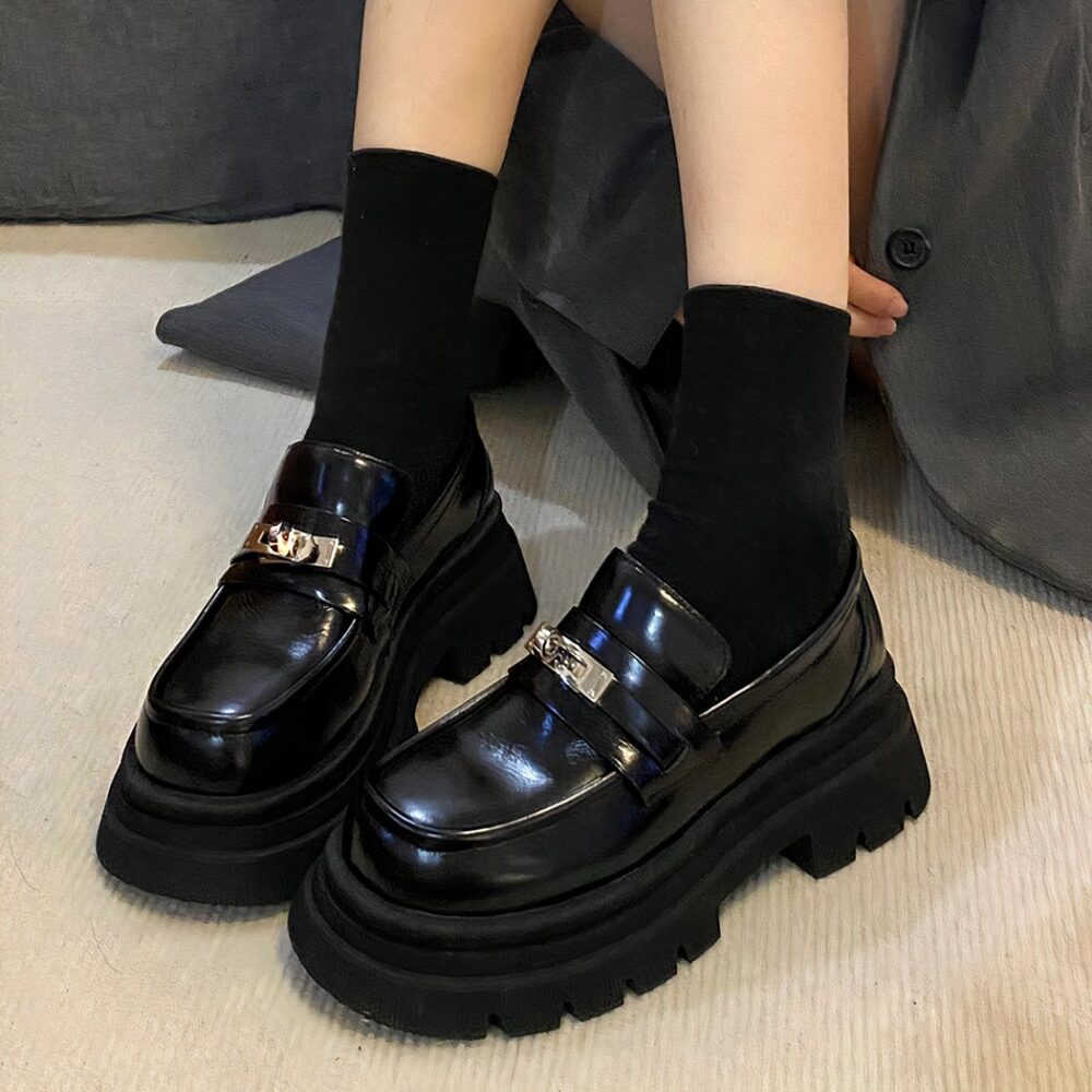 Black moccasins in shiny smooth leather with a thick sole at the feet of a woman sitting on the floor wearing short black socks.