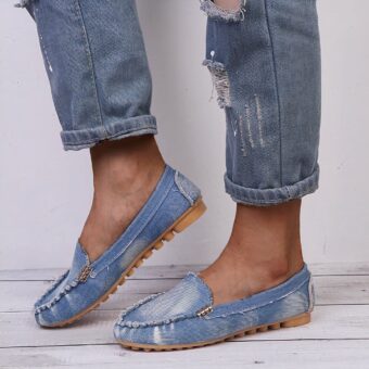 Pair of soft moccasins in faded blue denim with beige rubber soles with studs worn barefoot by a woman in blue ripped jeans with exposed ankles
