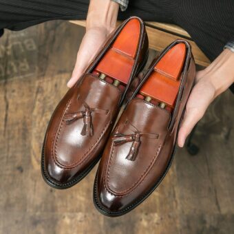 Dark, pointed-toe brown leather loafers with decorative tassels in the hands of a man holding them above a wooden floor.