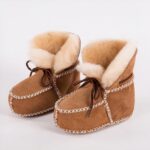 In profile, a pair of Indian moccasin booties in camel croute leather with white topstitching and exposed beige fur on the inside. Brown laces on top