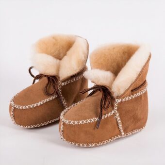 In profile, a pair of Indian moccasin booties in camel croute leather with white topstitching and exposed beige fur on the inside. Brown laces on top