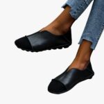 Pair of soft moccasins with round toe in black suede and smooth leather with exposed stitching worn with jeans on a light grey tiled floor