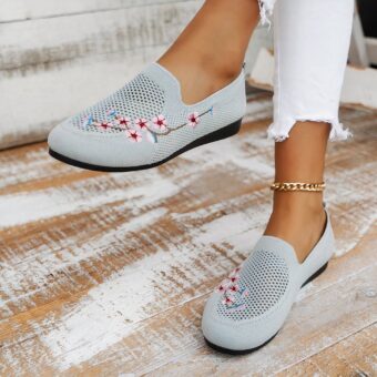 Gray openwork loafers with embroidered cherry blossom flowers and black soles. Worn with white jeans, a gold ankle chain on a bleached beige parquet floor.