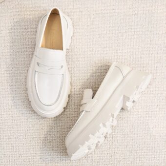 A pair of simple white smooth leather loafers with thick white soles set on a light beige ground.