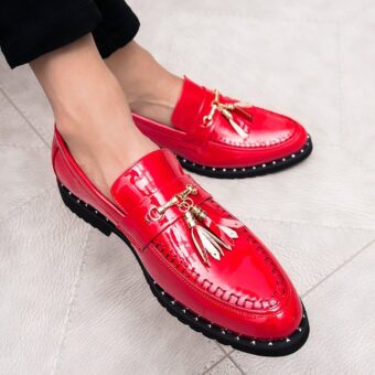 Bright red patent loafers with decorative tassels on the feet of a man sitting cross-legged in black suit pants.