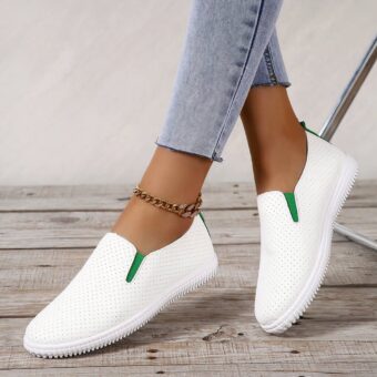 On a gray parquet floor, a pair of perforated white moccasins with small green elastics on the sides. They are worn barefoot with light blue jeans and ankle chain.