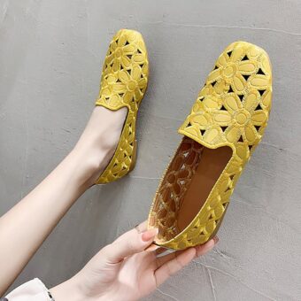 Pair of yellow openwork flat moccasins with flower motifs. One shoe is in a foot resting against a gray wall and the other is held in one hand