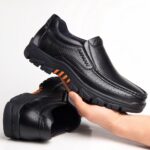 Photo of a pair of black leather orthopedic moccasins with thick soles, one shoe lying on the floor, the other held in one hand.