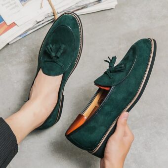 Emerald green suede loafers with decorative tassels on the foot to in the hand of someone sitting on a light gray floor.