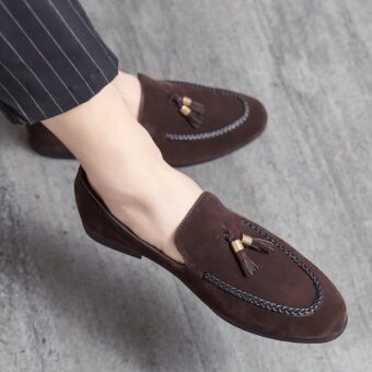 Suede loafers with decorative tassels and a leather twist on the seam, at the feet of a cross-legged man wearing black suit pants with thin white stripes.